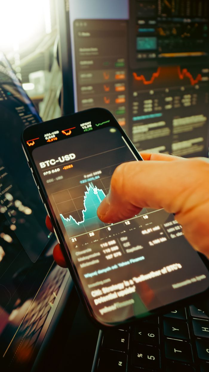 A hand holds a smartphone showing a BTC-USD chart amid digital trading screens, emphasizing crypto finance.