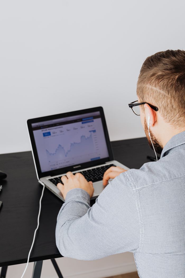 A man is using a laptop indoors to examine a stock market trends graph shown on the screen.